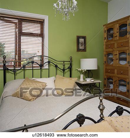 Stock Image - Traditional black metal bed in front of window with slatted wooden blind in small green bedroom. Fotosearch