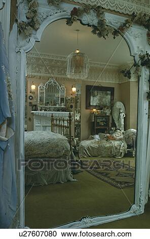 Stock Image - View through ornate white Victorian mirror of Victorian style bedroom with lace decorating walls and bed. Fotosearch