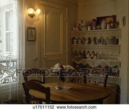Stock Image - Wall lights above table and chairs in small French dining room with lace-trimmed shelves. Fotosearch