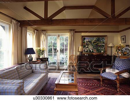Stock Image - White and yellow check sofa and glass-top table in livingroom extension with exposed beams and French windows . Fotosearch