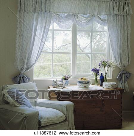 Stock Image - White armchair beside window with blue spotted voile curtains. Fotosearch