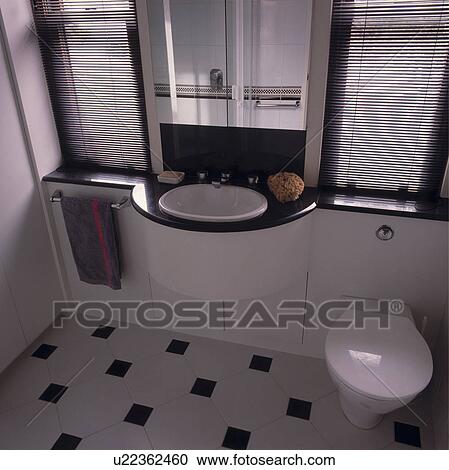 Stock Image - White basin in black+white fitted unit between black slatted blinds on windows in bathroom with black+white floor tiles. Fotosearch