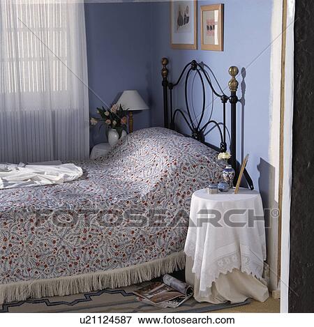 Stock Photo - White cloth on table beside wrought iron bed with patterned cover in small blue bedroom with white voile curtains. Fotosearch