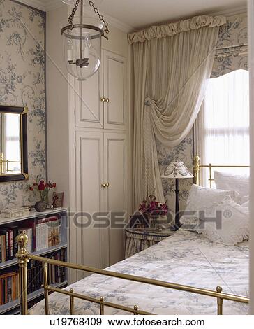 White Curtains And Blue White Blind Above Brass Bed In Bedroom With Fitted White Wardrobe And Blue Floral Wallpaper Stock Photo
