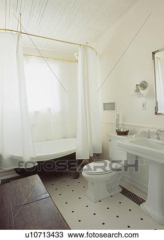Stock Image - White curtains around rolltop bath with black underside in white bathroom with pedestal basin and white and black floor. Fotosearch