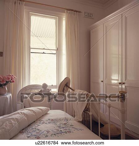 Picture - White curtains at window beside large white wardrobe in traditional bedroom with patterned white duvet on brass bed. Fotosearch