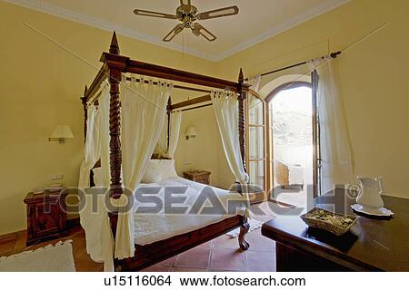 White drapes and linen on fourposter bed in pale yellow Spanish country bedroom with French doors open to patio View Large Photo Image Picture - White drapes and linen on fourposter bed in pale yellow Spanish country bedroom with French doors open to patio. Fotosearch