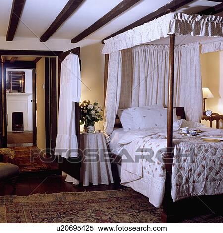Stock Photography - White drapes on antique fourposter bed with floral quilt in cottage bedroom with beamed ceiling. Fotosearch