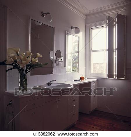 Stock Image - White lilies in vase on fitted vanity unit with basin in modern white bathroom with folding white shutters at window. Fotosearch