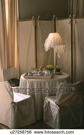 Stock Photograph - White loosecovers on chairs around table with circular cloth in dining room with white fabric and wood screen. Fotosearch
