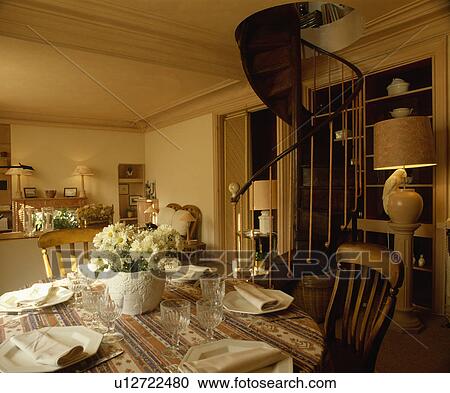 Stock Image - White plates and napkins on patterned cloth in eighties dining room with lighted lamps and spiral staircase. Fotosearch