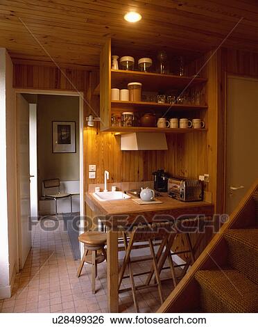 Stock Photograph - White sink in table and wooden wall shelves in compact wooden kitchen beside staircase. Fotosearch