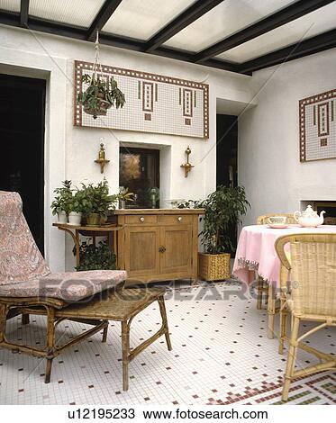 Stock Image - Wicker chaiselongue in dining room extension with pine cupboard and checked tiled flooring . Fotosearch