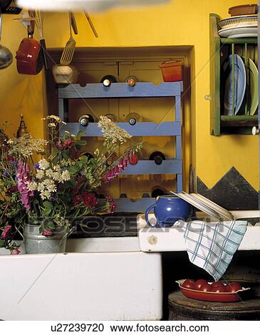 Stock Image - Wine bottles on blue wooden shelves in alcove above white ceramic sink with fresh flowers in metal vase. Fotosearch