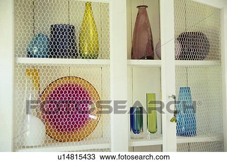 Stock Image - Wire doors open on colourful glass vases and plate on white shelves. Fotosearch