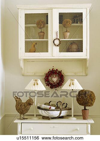 Wirework on doors of small wall cupboard above white lamps and dried moss tree with trug and wreath on chest of drawers View Large Photo Image Stock Photograph - Wirework on doors of small wall cupboard above white lamps and dried moss tree with trug and wreath on chest of drawers. Fotosearch