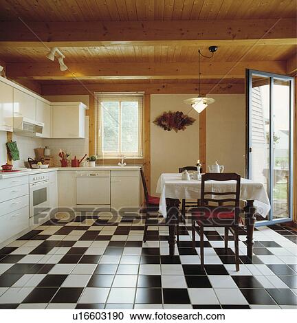 Stock Image - Wooden ceiling and black+white ceramic tiles floor in modern white country kitchen with traditional table and chairs. Fotosearch