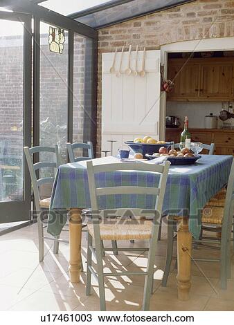Stock Image - Wooden chairs and blue tablecloth in dining room in conservatory extension. Fotosearch