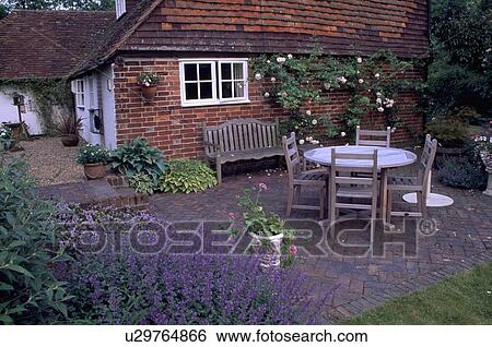 Stock Photograph - Wooden chairs and table on brick patio in front of country cottage. Fotosearch