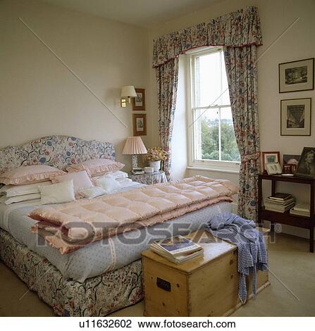 Stock Image - Wooden chest below upholstered bed with folded pink quilt in bedroom with floral curtains. Fotosearch