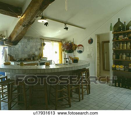 Stock Image - Wooden stools at white tiled worktop in in white country kitchen with large ceiling beam and white tiled floor. Fotosearch