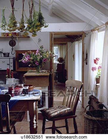 Stock Image - Wooden table and chairs below herbs drying on apex ceiling in white cottage kitchen with doorway to hall. Fotosearch