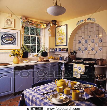 Stock Image - Yellow crockery on blue+white checked cloth in country kitchen with black Aga and painted blue fitted cupboards. Fotosearch