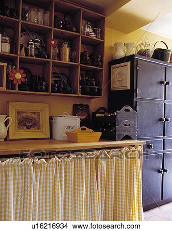 Picture - Yellow+white checked curtain below worktop in cottage kitchen with wooden cube wall-shelves. Fotosearch