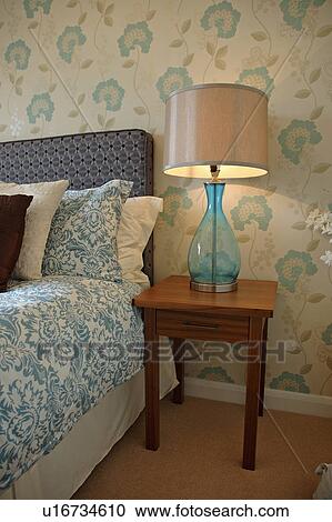 現代 青 Flower Patterned 壁紙 中に 寝室 で 青いガラス ランプ 上に 木 ベッドサイド テーブル と青 そして 白 シーツと枕カバー ストックイメージ U Fotosearch