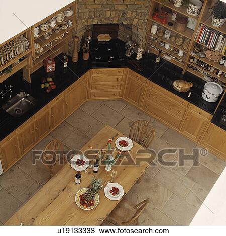 Stock Image - Aerial view of long pine table in country kitchen with black granite worktops on wooden fitted cupboards. Fotosearch
