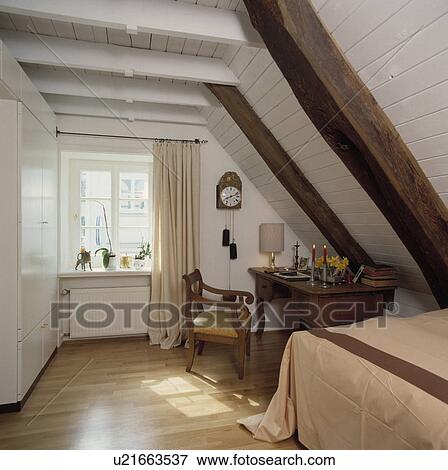 Stock Photo - Antique desk and chair below sloping white wooden ceiling with large beams in attic bedroom with wooden flooring. Fotosearch