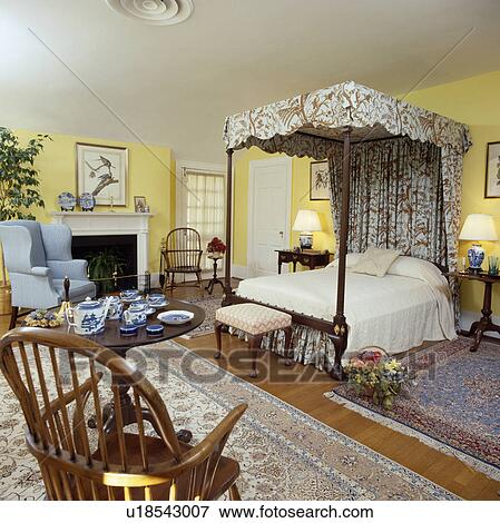 Stock Photo - Antique four-poster bed and large oriental rugs in yellow traditional bedwoorm with blue armchair beside fireplace. Fotosearch