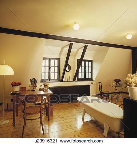 Stock Image - Antique pine table and chair in large attic bathroom with freestanding bath and wooden flooring. Fotosearch