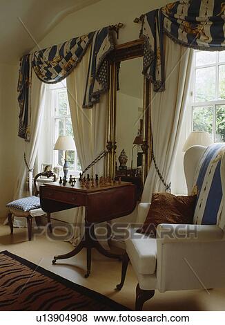 Antique table between windows with swagged curtains in traditional living room View Large Photo Image Stock Photo - Antique table between windows with swagged curtains in traditional living room. Fotosearch