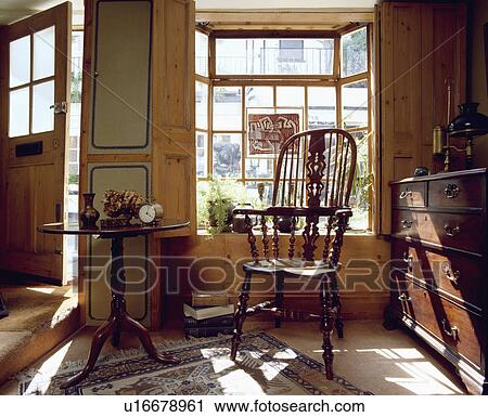 Stock Image - Antique Windsor chair and circular Victorian table in front of window in hall with chest of drawers. Fotosearch
