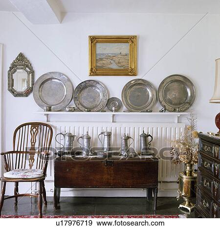 Stock Photo - Antique Windsor chair and oak chest with pewter jugs below collection of pewter plates on shelf above radiator. Fotosearch