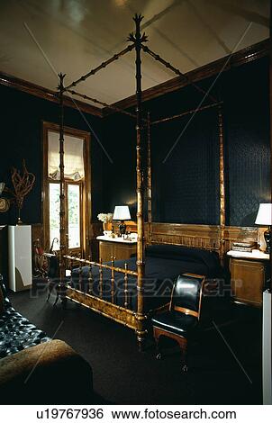 Stock Photograph - Bamboo fourposter bed with black linen in masculine black bedroom with lighted lamps on either side of the bed. Fotosearch