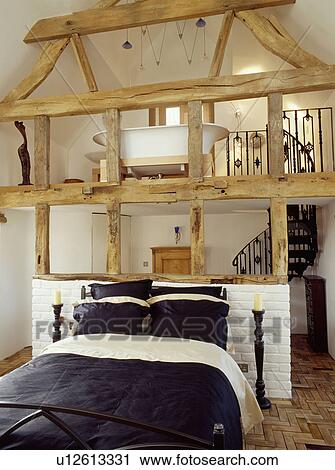 Stock Image - Bed with black+white linen in bedroom with plaster removed from beamed wall below mezzanine bathroom. Fotosearch