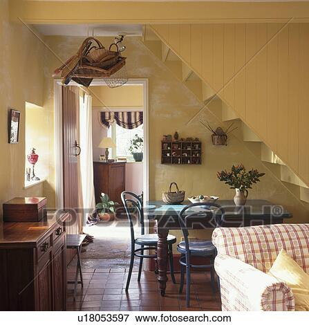 Stock Photo - Bentwood chairs and wooden table below staircase in yellow cottage kitchen with terracotta tiled floor and red check sofa. Fotosearch
