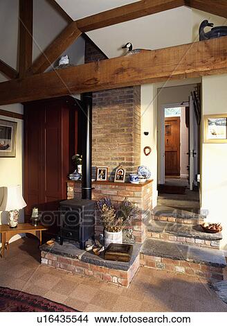 Picture - Black cast-iron stove in cottage hall with beams and exposed brick wall and steps up to open door. Fotosearch