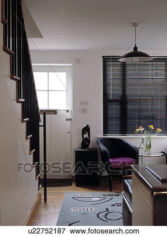Stock Photo - Black Lloyd Loom chair below black slatted blind in small modern living room with staircase. Fotosearch