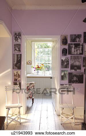 Stock Image - Black+white photographs on wall of pink landing with white chairs and floorboards and door open to bathroom. Fotosearch