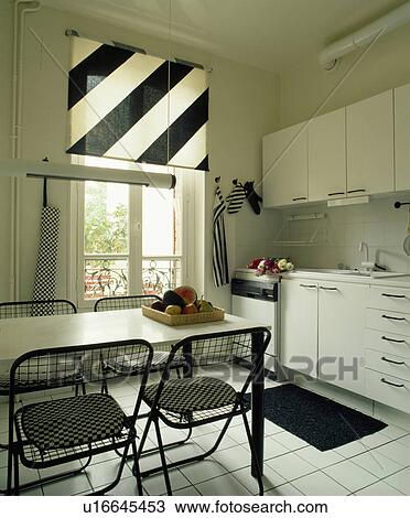 Stock Image - Black+white striped blind on window in modern white kitchen with white table and black metal chairs. Fotosearch