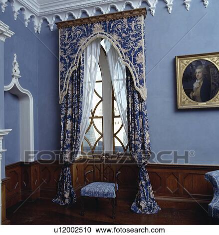 Stock Image - Blue+white patterned curtains and pelmet on Gothic window in blue room with white Gothic cornice. Fotosearch