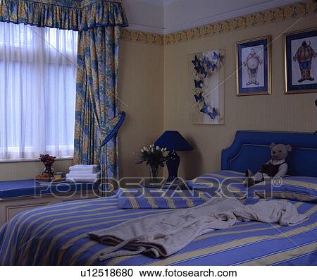 Stock Image - Blue+yellow striped quilt and pillows on bed in neutral bedroom with white net and blue+yellow floral curtains. Fotosearch