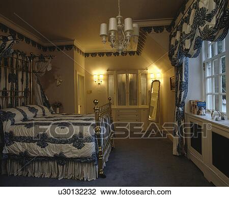 Brass half-tester bed with blue+white ribbon pattern bedcover and matching curtains at window of traditional bedroom View Large Photo Image Stock Image - Brass half-tester bed with blue+white ribbon pattern bedcover and matching curtains at window of traditional bedroom. Fotosearch