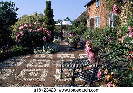Stock Image - Brick and pebble paving in front of cottage with pink roses in large garden in summer. Fotosearch