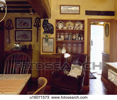 Stock Image - Brown leather armchair in front of alcove shelves with glass jars and pottery in yellow cottage kitchen . Fotosearch