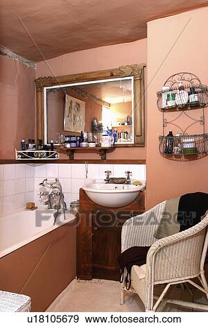 Stock Photo - Carved antique mirror above basin and bath in pastel pink cottage bathroom with cream Lloyd Loom chair. Fotosearch