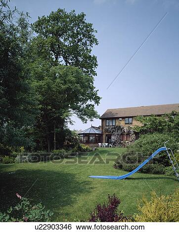 Stock Photograph - Childrens blue slide on lawn in country garden with tall trees and house in the background. Fotosearch